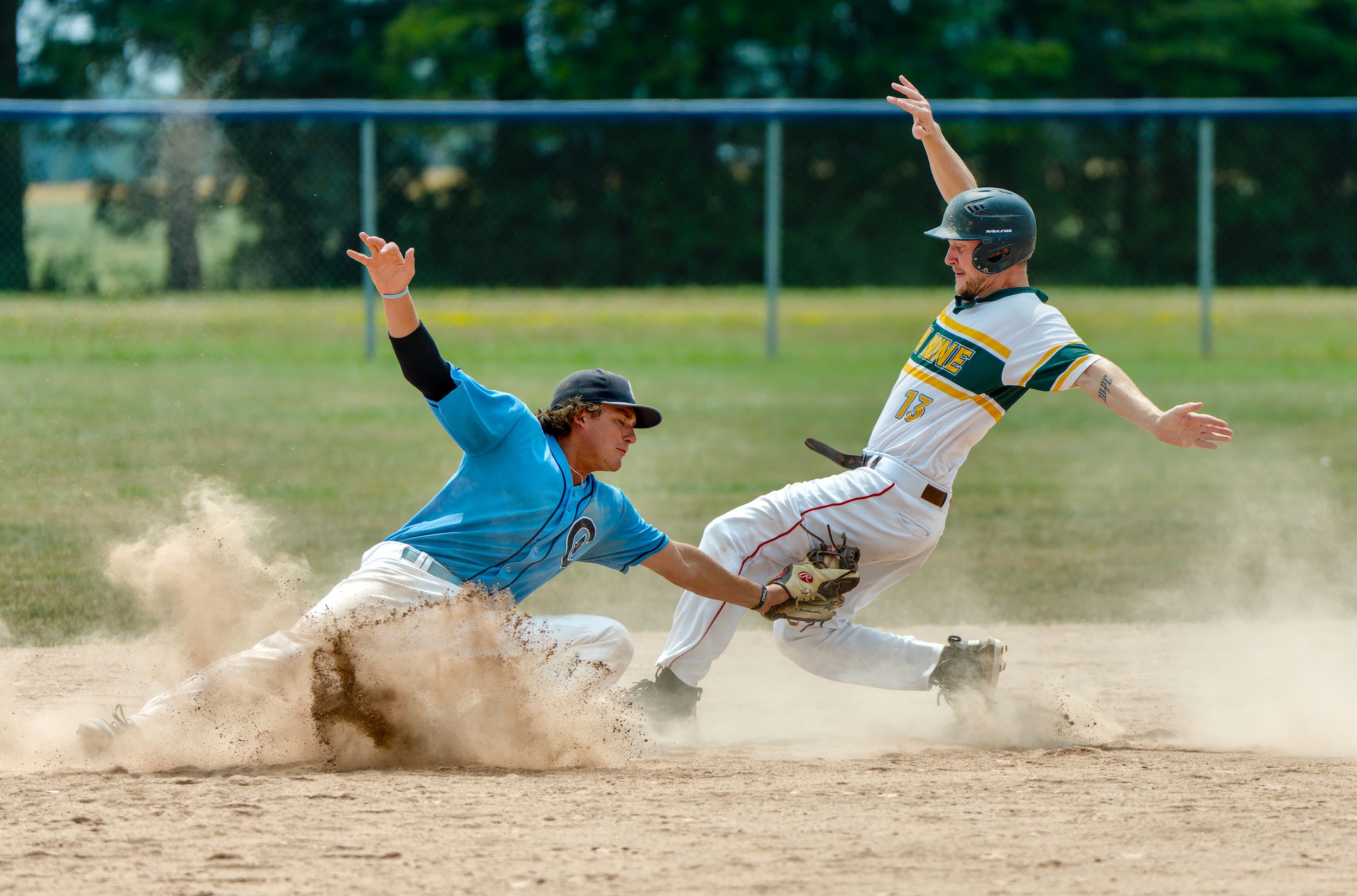 A fielder in blue lunges to tag a runner sliding into second base through an exploding cloud of infield dust, the runner's arms thrown wide.
